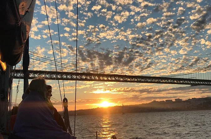 Guests wrapped in blankets on a sailboat as the sun sets under the 25 de Abril bridge